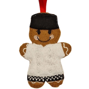 May include: A gingerbread man Christmas decoration. The brown felt ornament has a black hat and a white chef's coat. The coat's hem features a black and white chequered pattern. A red ribbon is attached for hanging.