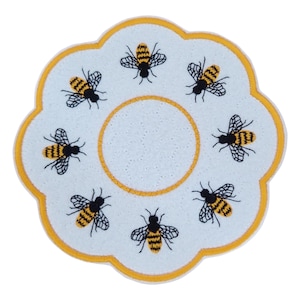 May include: A white, flower-shaped decorative mat with a yellow border. The mat features a central circle and is adorned with seven embroidered bees in black and yellow, creating a charming and whimsical design.