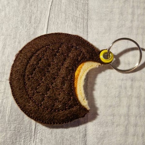 May include: A brown felt keyring in the shape of a cookie with a bite taken out. The cookie features a quilted pattern and a yellow and white filling. It has a silver ring and a yellow grommet.