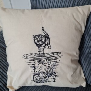 Peut inclure: Coussin carré crème avec un motif brodé d'un chaton se tenant dans l'eau, avec son reflet en dessous. Le chaton est gris et noir, et l'eau est représentée par des cercles concentriques.