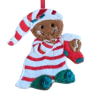 Könnte beinhalten: Ein Weihnachtsornament aus Filz, das einen Lebkuchenmann darstellt. Das Ornament ist braun mit einer weiß-rot gestreiften Nachtmütze und Pyjama. Der Lebkuchenmann hält einen roten Strumpf mit einem Lebkuchen. Das Ornament hat ein rotes Band zum Aufhängen.