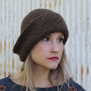 May include: A woman wearing a brown felt cloche hat with a slightly slouched crown. She has blonde hair and is wearing a dark blue floral print blouse.