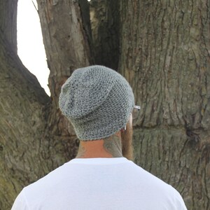 May include: A gray knitted beanie with a slouchy fit. The beanie is worn by a person with a beard and tattoos.