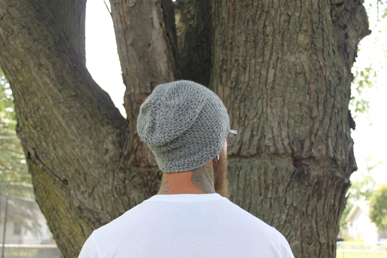May include: A gray knitted beanie with a slouchy fit. The beanie is worn by a person with a beard and tattoos.