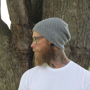 May include: A gray knitted beanie with a slouchy fit. The beanie is worn by a person with a beard and glasses. The person is standing in front of a tree.