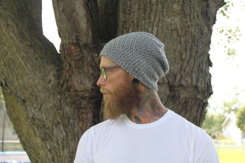 May include: A gray knitted beanie with a slouchy fit. The beanie is worn by a person with a beard and glasses. The person is standing in front of a tree.