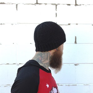 May include: A black knitted beanie with a ribbed texture. The beanie is being worn by a person with a beard and a tattoo on their neck.