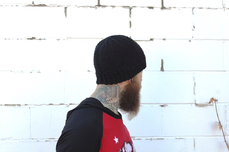 May include: A black knitted beanie with a ribbed texture. The beanie is being worn by a person with a beard and a tattoo on their neck.