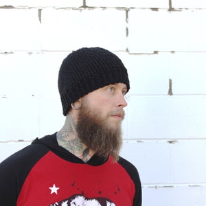 May include: A black knitted beanie with a folded brim. The beanie is being worn by a person with a beard and tattoos.