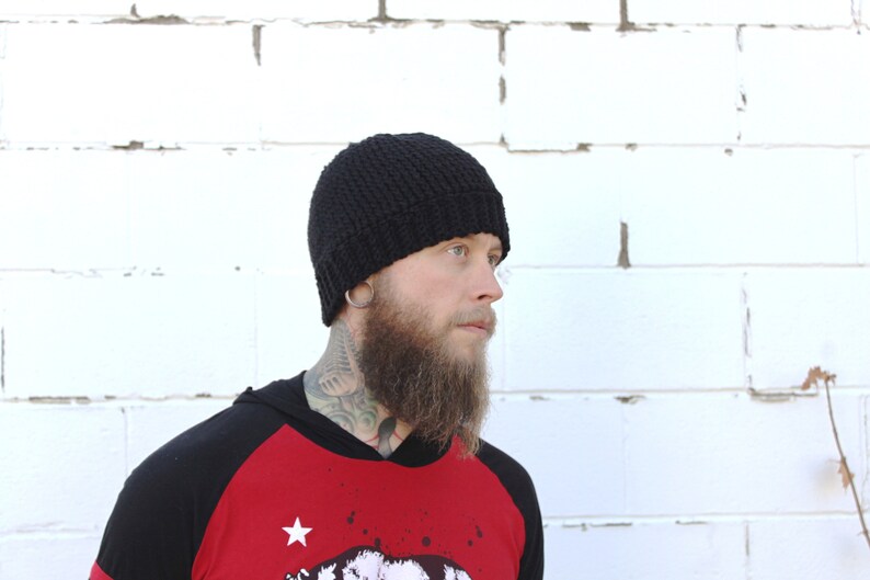 May include: A black knitted beanie with a folded brim. The beanie is being worn by a person with a beard and tattoos.