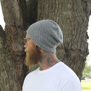 May include: A gray slouchy beanie hat with a textured knit pattern. The hat is worn by a person with a beard and tattoos.