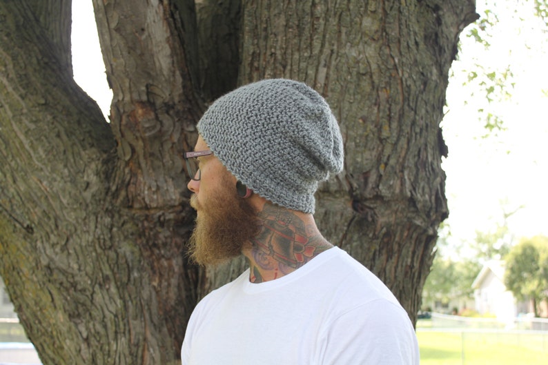 May include: A gray slouchy beanie hat with a textured knit pattern. The hat is worn by a person with a beard and tattoos.