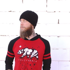 May include: A man wearing a black beanie, a red and black baseball shirt with a white bear graphic and the text "CALIFORNIA", and a long beard.