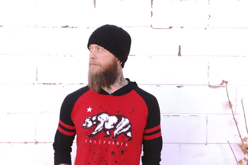 May include: A man wearing a black beanie, a red and black baseball shirt with a white bear graphic and the text "CALIFORNIA", and a long beard.