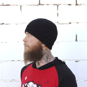 May include: A black knitted beanie with a slouchy fit. The beanie is being worn by a person with a beard and tattoos.