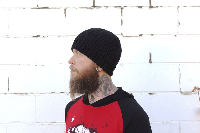 May include: A black knitted beanie with a slouchy fit. The beanie is being worn by a person with a beard and tattoos.