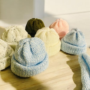 May include: A collection of handmade knit beanies in various colors, including light blue, cream, pink, and olive green. The hats are arranged on a wooden surface, showcasing their soft texture and simple design. These beanies are perfect for babies and toddlers.