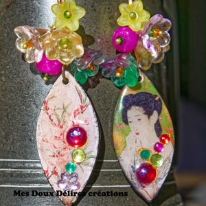 Op de afbeelding: Een paar hangende oorbellen met bladvormige hangers met afbeeldingen van een persoon en bloemmotieven. De oorbellen zijn versierd met kleurrijke gefacetteerde strass-steentjes en clusters van doorschijnende gele, roze en groene bloemkralen.