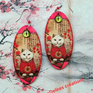 May include: A pair of oval earrings with a white cat in a red outfit, set against a backdrop of Japanese script and red flowers. The earrings have a bright pink border and a green eye-shaped bead at the top. The text "MesDouxDelires créations" is at the bottom.