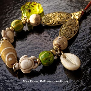 Può includere: Un bracciale con un mix di perline che include forme di tartaruga bianche, una rana in vetro verde, una perlina verde maculata, una perlina marrone a motivi e una conchiglia di ciprea bianca. Include accenti in metallo dorato e il testo Mes Doux Délires créations.