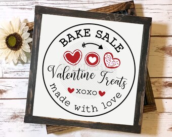 Op de afbeelding: Een wit bord met zwarte tekst en rode en roze harten met de tekst "Bake Sale Valentine Treats made with love xoxo".