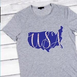 Op de afbeelding: Een lichtgrijs t-shirt met een blauwe omtrek van de Verenigde Staten en het woord "USA" in witte letters. Rode sterren zijn in de letters geplaatst. Het t-shirt heeft een ronde hals.