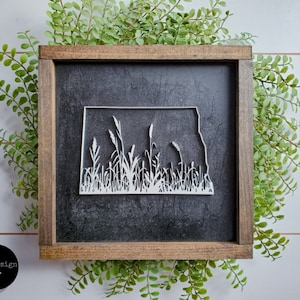May include: A black framed wood sign with a white silhouette of the state of North Dakota with grass growing inside the state outline.