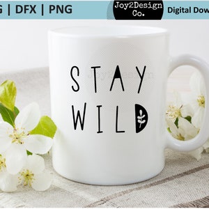 May include: White ceramic mug with black text that reads "Stay Wild" and a small leaf design. The mug is sitting on a light brown surface with white flowers.