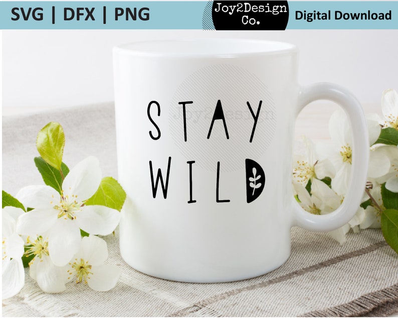 May include: White ceramic mug with black text that reads "Stay Wild" and a small leaf design. The mug is sitting on a light brown surface with white flowers.
