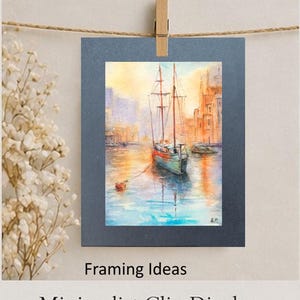 May include: A watercolor painting of a sailboat on a body of water, with buildings in the background. The painting is displayed on a dark blue mat, clipped to a clothesline with a wooden clip. The words "Framing Ideas" and "Minimalist Clip Display" are below the painting.