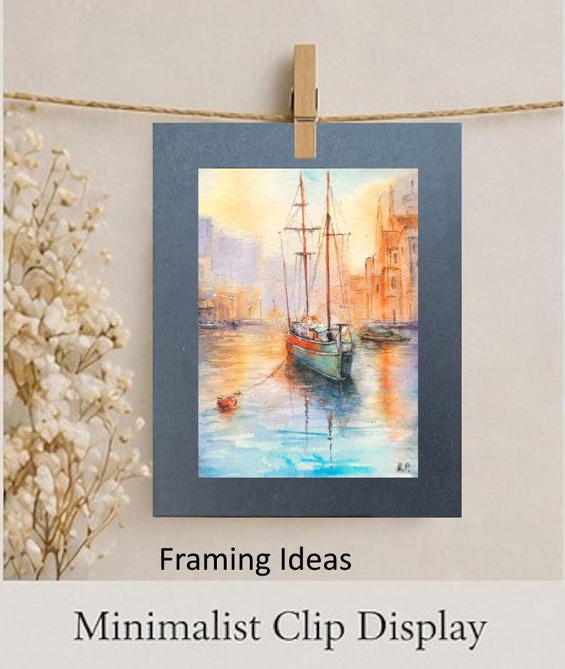 May include: A watercolor painting of a sailboat on a body of water, with buildings in the background. The painting is displayed on a dark blue mat, clipped to a clothesline with a wooden clip. The words "Framing Ideas" and "Minimalist Clip Display" are below the painting.