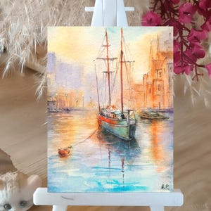 May include: Watercolor painting of a sailboat in a harbor, displayed on a small white easel. The artwork features a boat with red and brown masts, docked in calm water. Buildings line the background, and the sky is a blend of yellow and orange.
