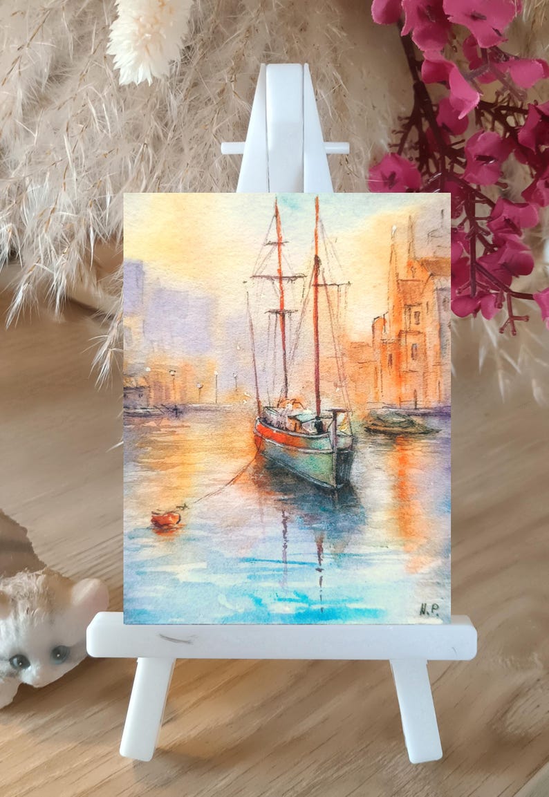 May include: Watercolor painting of a sailboat in a harbor, displayed on a small white easel. The artwork features a boat with red and brown masts, docked in calm water. Buildings line the background, and the sky is a blend of yellow and orange.