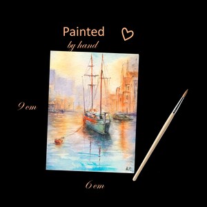 May include: Watercolor painting of a sailboat in a harbor, with buildings in the background. The painting is 9 cm x 6 cm and has the text "Painted by hand". A paintbrush is next to the painting.