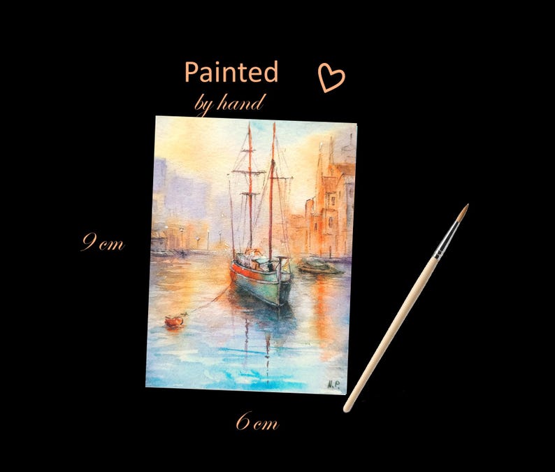 May include: Watercolor painting of a sailboat in a harbor, with buildings in the background. The painting is 9 cm x 6 cm and has the text "Painted by hand". A paintbrush is next to the painting.