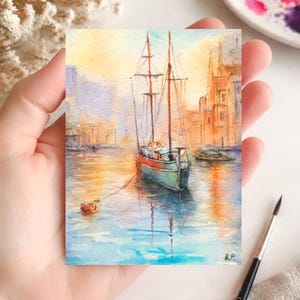 May include: Watercolor painting of a sailboat on the water. The boat has a red and green hull with two masts. The background features buildings and a sunset sky. The painting is on a small rectangular piece of paper.