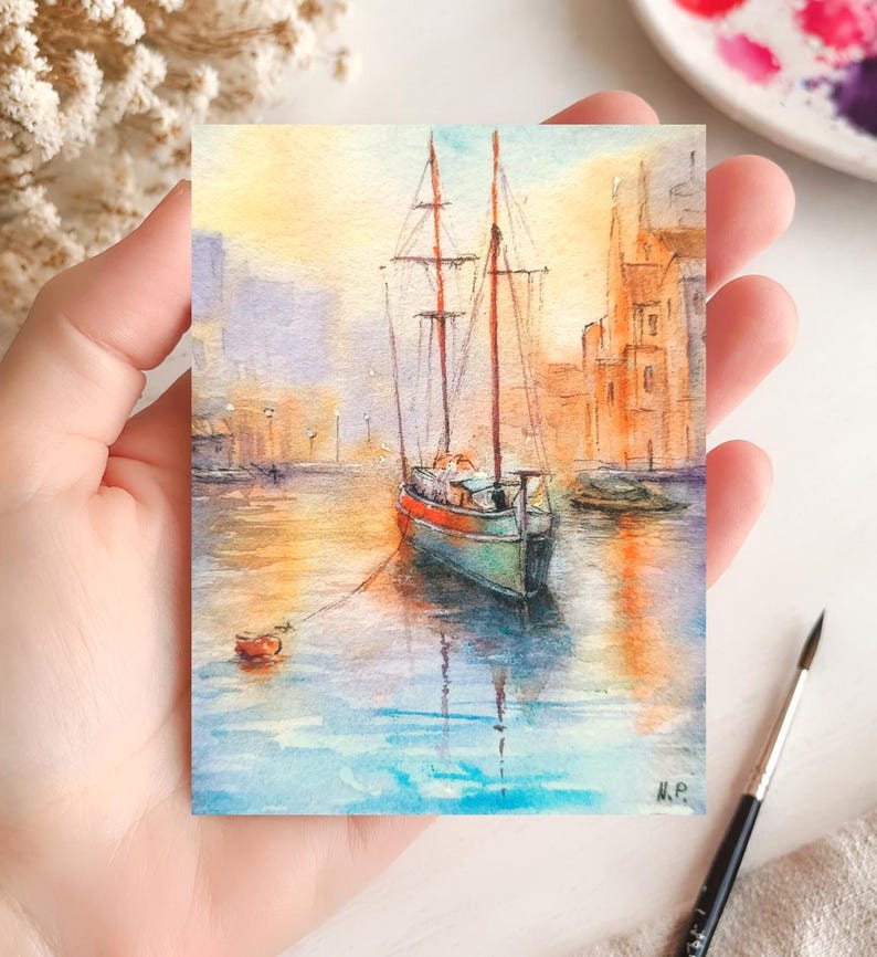 May include: Watercolor painting of a sailboat on the water. The boat has a red and green hull with two masts. The background features buildings and a sunset sky. The painting is on a small rectangular piece of paper.