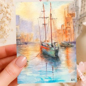 May include: Watercolor painting of a sailboat in a harbor. The boat has a red and green hull, with masts and rigging. The water reflects the warm colors of the sunset and the buildings in the background. The painting is signed "N.P."