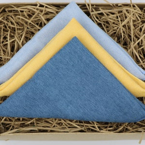 Puede incluir: Tres paños doblados en una caja llena de papel de embalaje marrón. Los paños son azules, amarillos y azul claro.