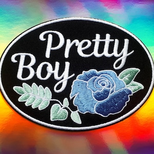 May include: An embroidered patch featuring the words "Pretty Boy" in white script within a black oval. A blue rose and green leaves are below the text. The background has a colourful, iridescent finish.
