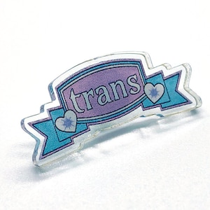 May include: A clear acrylic pin featuring the word "trans" in a purple banner. The banner is outlined in white and has blue ribbon-like accents with heart details. The pin is a symbol of transgender pride.
