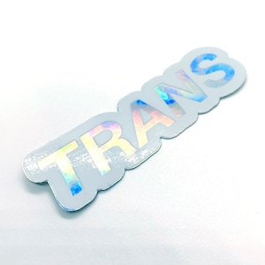 Puede incluir: Una pegatina holográfica con la palabra "TRANS" en una fuente estilizada. La pegatina tiene un borde azul claro y un acabado reflectante de color arcoíris. La pegatina está sobre un fondo blanco.