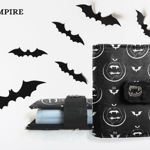 May include: Black wallet with a vampire themed design. The wallet has a black background with white vampire teeth and bats. The wallet is surrounded by black bats.