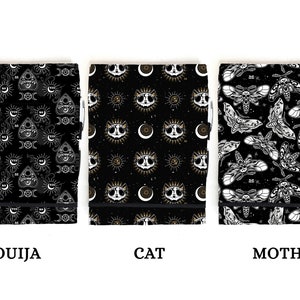 May include: Three black notebooks with different designs. The first notebook has a white and gold pattern of an Ouija board, moons, and suns. The second notebook has a white and gold pattern of a cat's face, moons, and suns. The third notebook has a white and gold pattern of moths.