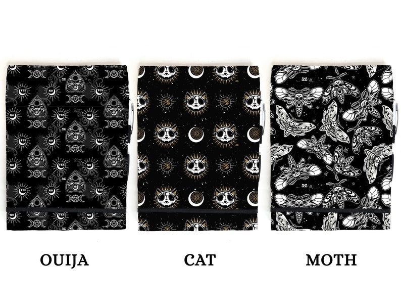 May include: Three black notebooks with different designs. The first notebook has a white and gold pattern of an Ouija board, moons, and suns. The second notebook has a white and gold pattern of a cat's face, moons, and suns. The third notebook has a white and gold pattern of moths.