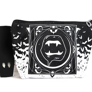 May include: Black and white cosmetic bag with a vampire-themed design. The bag features a graphic of vampire fangs and bats. A small black coffin-shaped item is next to the bag. The bag has a black zipper and a small black loop.