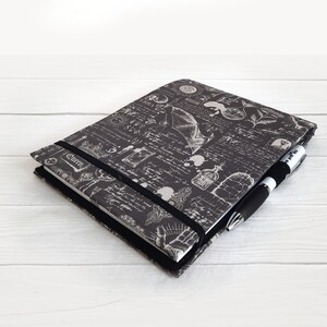 May include: A black and white journal with a gothic design featuring a bat, a skull, and other occult symbols. The journal has a black elastic closure and a black pen is included.