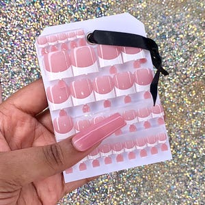 May include: A set of pink and white press-on nails with a French manicure design. The nails are arranged on a white card with a black ribbon.