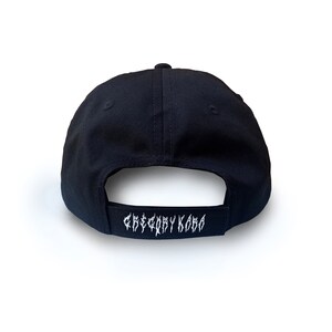 K&ouml;nnte beinhalten: Schwarze Baseballkappe mit einem wei&szlig;en gestickten Logo, das auf der R&uuml;ckseite "Gregory Kara" lautet.