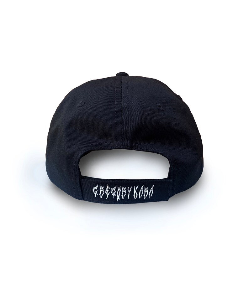 K&ouml;nnte beinhalten: Schwarze Baseballkappe mit einem wei&szlig;en gestickten Logo, das auf der R&uuml;ckseite "Gregory Kara" lautet.